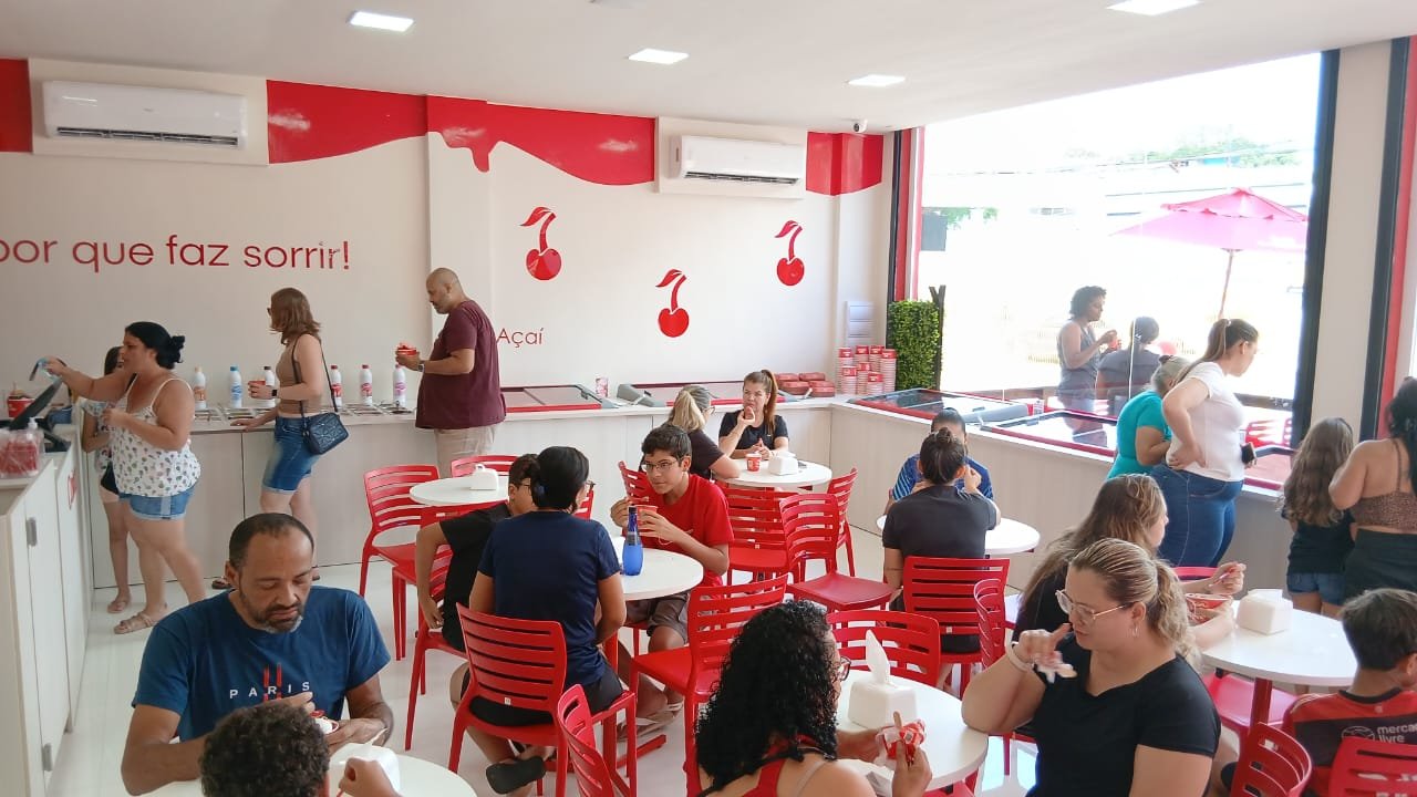 Interior of a 60 Sabores unit with customers enjoying açaís, red chairs, thematic decoration with hanging red cherries, service counter, cozy and well-lit franchise environment